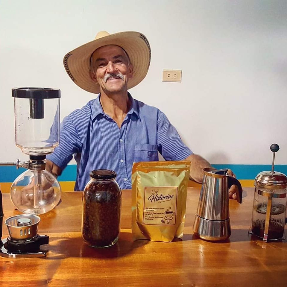 A smiling coffee farmer in a sombrero behind a table of siphon, moka pot, French press and his own bagged coffee