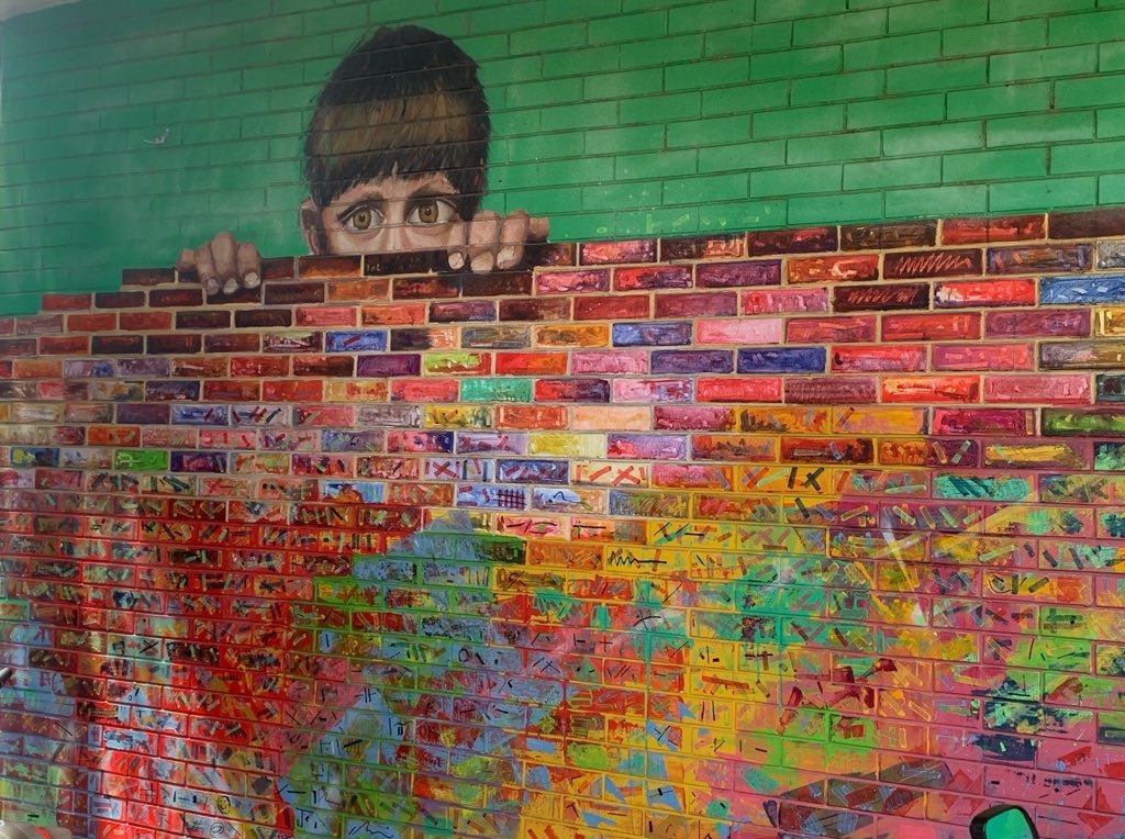 A mural of a boy peering over a wall of hand-painted, multicolored bricks