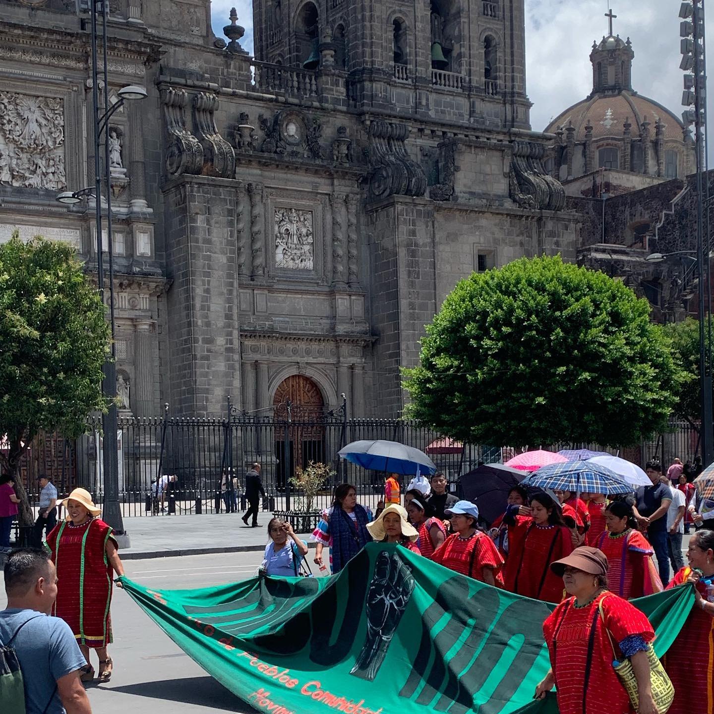 Indigenous women in red huipiles carry a long green banner past the Metropolitan Cathedral in the Zócalo