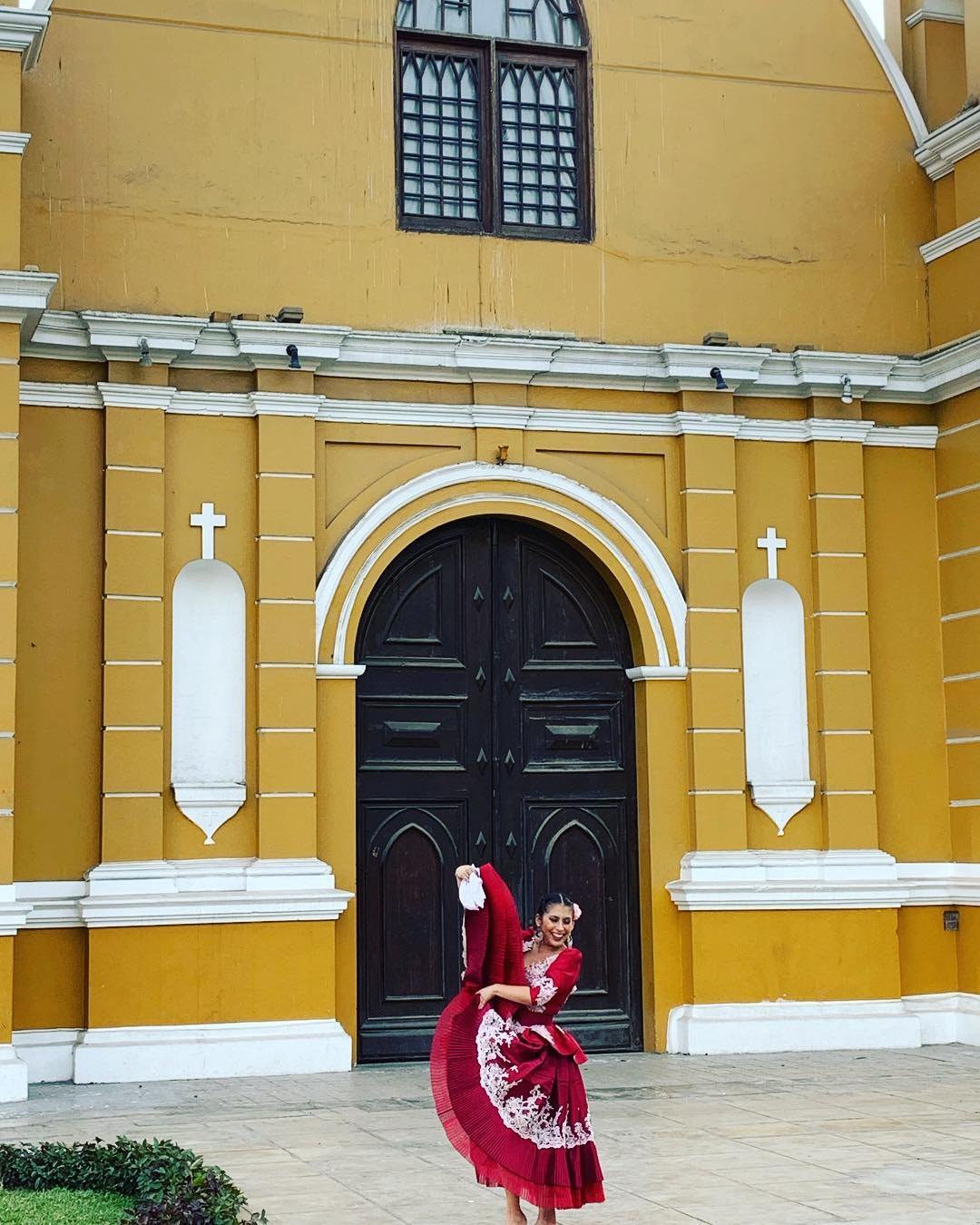 A marinera dancer in a red dress, mid-turn, in front of a yellow church
